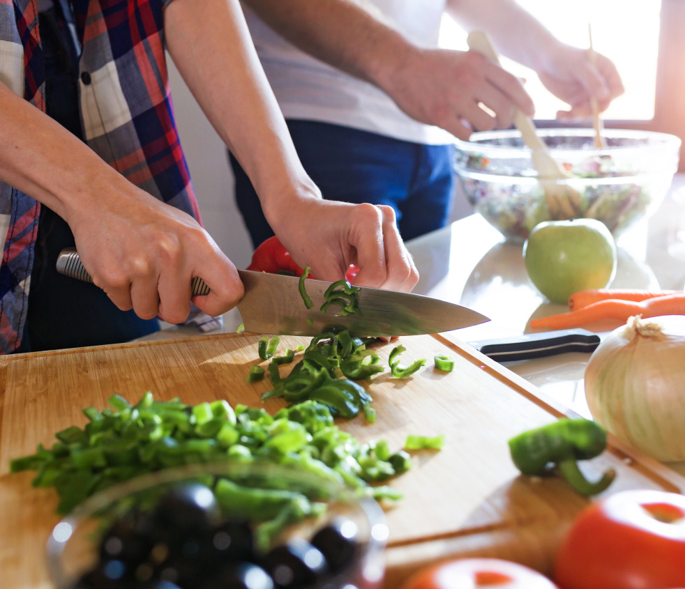 Zwei junge Männer bereiten ein Essen vor. Sie schneiden Gemüse und rühren Salat an. Es sind nur die Hände und ein Teil der Arbeitsplatte zu sehen.